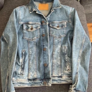Klassisk blå jeansjacka - Säljer en klassisk blå jeansjacka med knappar framtill och två bröstfickor med lock. Jackan har långa ärmar och raka sömmar för en tidlös look. Perfekt att slänga på sig över en hoodie eller t-shirt.
