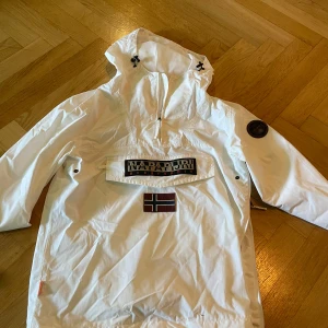 Vit vindjacka från Napapijri - Säljer en vit vindjacka från Napapijri med stor huva och dragkedja vid halsen. Jackan har en stor ficka framtill med logotyp och norsk flagga, samt en patch på ärmen. Perfekt för blåsiga dagar.