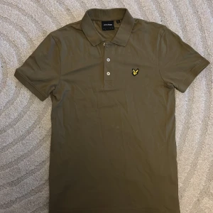 Olivgrön pikétröja från Lyle & Scott - Säljer en olivgrön piké från Lyle & Scott med klassisk krage, knappar och den ikoniska broderade gula örnen på bröstet. Perfekt för en stilren och avslappnad look. Använd fåtals gånger. Finns en liten fläk( bild 4) syns knappast men är fortfarande där!