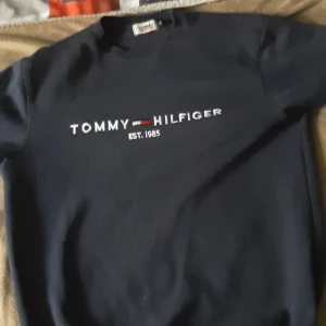 Mörkblå Tommy Hilfiger tröja - Snygg mörkblå tröja från Tommy Hilfiger med klassisk logga och texten 'EST. 1985' broderad på bröstet. Tröjan har rund halsringning och långa ärmar, perfekt för en stilren och avslappnad look.Har storlek M