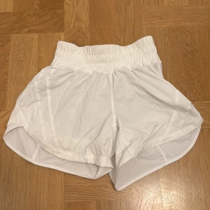 Vita träningsshorts från Lululemon - Snygga vita träningsshorts från Lululemon med bred resår i midjan och rundade benslut. Lätta och luftiga i syntetmaterial, perfekta för löpning eller gym. Klassisk Lululemon-logga på benet och innerbyxa för extra komfort.