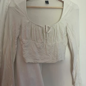 Vit långärmad croppad topp från SHEIN - Säljer en vit långärmad croppad topp från SHEIN med knyt framtill och lätt rynkade detaljer vid bysten. Toppen har en rundad urringning och är tillverkad i ett mjukt bomullsmaterial. 