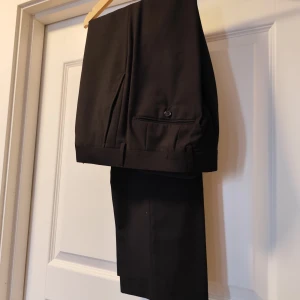 Svarta kostymbyxor från Dressmann - Klassiska svarta kostymbyxor från Dressmann med pressveck och diskreta sidofickor. Byxorna har hällor för bälte, stängs med hake och dragkedja och är tillverkade i en blandning av polyester och viskos för en mjuk och följsam känsla. Använt en gång