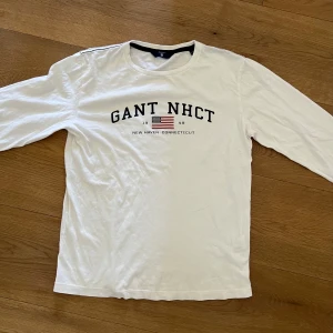 Vit långärmad tröja från GANT - Säljer en vit långärmad tröja från GANT med tryck 'GANT NHCT' och amerikansk flagga på bröstet. Tröjan har rund halsringning och klassisk passform. Perfekt för en stilren och avslappnad look.