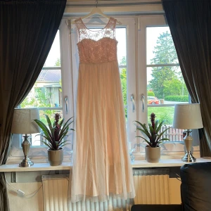 Rosa långklänning med spetsdetaljer - Elegant långklänning i rosa färg med vacker spets upptill och transparent mesh vid axlarna. Kjolen är i ett lätt, flytande material som ger en romantisk känsla. Perfekt för speciella tillfällen.