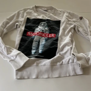 Vit Moncler sweatshirt med astronautprint - Vit sweatshirt från Moncler med stort tryck av en astronaut och texten 'MOONCLER' på bröstet. Tröjan har långa ärmar, ribbade muddar och klassisk Moncler-logga på ärmen. Perfekt för dig som gillar streetstyle och unika prints.