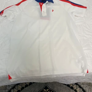 Vit pikétröja Peak Performance M - Snygg vit pikétröja från Peak Performance med blå och röda detaljer på krage och axlar. Klassisk pikéknäppning framtill och logga på bröstet. Perfekt för en sportig look, tillverkad i bomull för skön känsla. R&D-detalj bak på nederkanten.