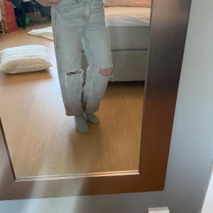 Ljusgrå wide leg jeans med slitningar - Säljer ett par ljusgrå jeans med wide leg-modell och slitna detaljer över knäna. Byxorna har klassisk femficksdesign och hög midja. Perfekta för en avslappnad och trendig look.
