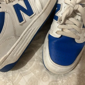 Vita och blå New Balance sneakers - Säljer ett par vita och blå New Balance sneakers med klassisk design. Skorna har snörning, rund tå och platt sula. Perfekta för dig som gillar sportig stil och vill ha bekväma skor till vardags.