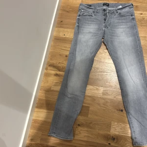 Ljusgrå jeansbyxor från Jack & Jones - Säljer ett par ljusgrå jeans från Nudie Jeans med klassisk femficksdesign och knappgylf. Jeansen har raka ben och diskreta sömmar på bakfickorna. Perfekta för en avslappnad stil.  Slim fit.