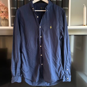 Mörkblå skjorta från Ralph Lauren - Säljer en mörkblå långärmad skjorta från Ralph Lauren i slim fit-modell. Skjortan har klassisk krage, knappar framtill och den ikoniska broderade loggan i gult på bröstet. Tillverkad i lätt bomullstyg. Har även Qr kod som såklart funkar och är i riktigt bra skick! Skriv för frågor och funderingar 