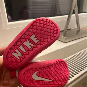 Vita och rosa Nike barnskor - Söta små Nike barnskor i vitt och rosa med kardborreband för enkel på- och avtagning. Skorna har en mjuk och bekväm insida med en slitstark sula. Perfekta för små fötter som är på språng.