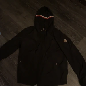  Moncler windbreaker  - Säljer en stilren svart jacka från Moncler med huva och dragkedja. Jackan har en broderad logotyp på ärmen och knappar framtill. Perfekt nu till våren när de blir blosiga men varmare dagar. Snörena borttagna då ena metall grejen gick av så tog bort dem då helt. Skriv vid funderingar;), obs pris ej hugget i sten kan sänkas vid snabb affär.