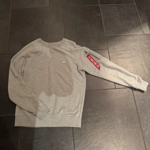 Grå tröja från Alpha Industries - Säljer en grå tröja från Alpha Industries. Tröjan har en liten ficka med dragkedja och en röd tagg med texten 'Remove Before Flight'. Använd max 10 gånger. Tyvärr inget kvitto. 