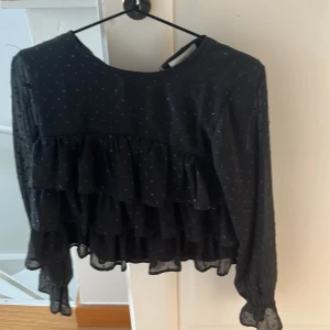 Svart blus från Gina Tricot - Snygg svart blus från Gina Tricot med volanger och små prickar. Blusen har långa ärmar och en luftig passform, perfekt för både vardag och fest. Den är gjord i ett lätt och bekvämt material.