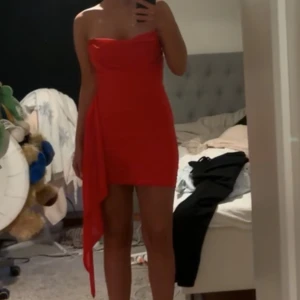 Röd axelbandslös klänning - Snygg röd axelbandslös klänning med draperad design och asymmetrisk detalj på sidan. Perfekt för en kväll ute eller speciella tillfällen. Klänningen har en tight passform som framhäver figuren.