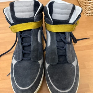 Grå och gula sneakers från Louis Vuitton - Snygga grå sneakers med gula detaljer från Louis Vuitton. Skorna har en unik design med snörning och ett gult spänne för extra stil. Sulan är vit med LV-logotypen i gult. Perfekta för den som vill ha något exklusivt och trendigt.