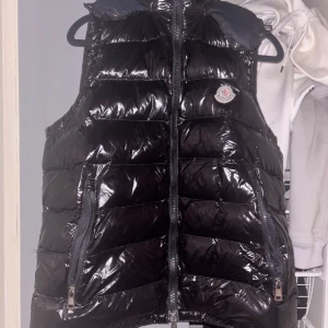 Svart dunväst från Moncler - Snygg svart dunväst från Moncler med glansig yta och dragkedja framtill. Västen har en klassisk design med Moncler-logga på bröstet. Perfekt för att hålla värmen med stil.pris kan diskuteras vid snabb affär