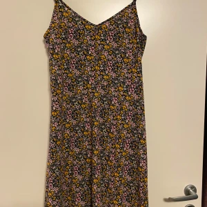 Blommig klänning från Vero Moda - Säljer en söt blommig klänning från Vero Moda i storlek S. Klänningen har ett färgglatt mönster med rosa, gula och vita blommor på en svart bakgrund. Mycket tunt och svalt material, perfekt för varma dagar! 