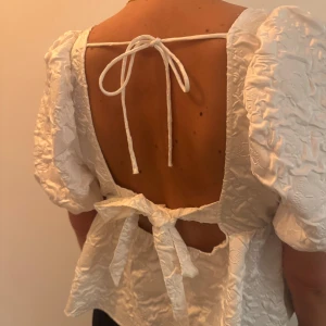 Vit blus med puffärmar - Säljer en söt vit blus med puffiga kortärmar och knytband i ryggen. Blusen har en fyrkantig urringning och ett fint texturerat mönster. Perfekt  till sommarn.