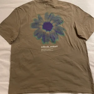 Beige t-shirt med blomtryck från H&M - Snygg beige t-shirt från H&M med ett stort blommönster i lila och blått på baksidan. 