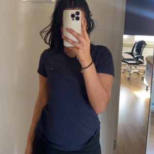 Mörkblå t-shirt från Tommy Hilfiger - Säljer en stilren mörkblå t-shirt från Tommy Hilfiger med deras ikoniska logga på bröstet. T-shirten är kortärmad och gjord i mjuk bomull, perfekt för en avslappnad look. Passar bra till jeans eller shorts! Är Xxs i storlek men passar mig som har xs. Har använt 1-2 gånger. 