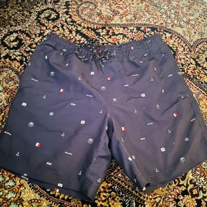 Mönstrade badshorts från DeFacto - Snygga mörkblå badshorts från DeFacto med ett marint mönster av små symboler som ankare och flaggor. Shortsen har snörning i midjan och fickor på sidor och är tillverkade i ett lätt och bekvämt material med innerfoder i mesh.
