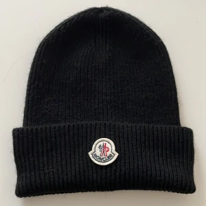 Svart mössa från Moncler - Snygg svart stickad mössa från Moncler med broderad logotyp framtill. Perfekt för att hålla värmen under kyliga dagar. Klassisk design med uppvikt kant.