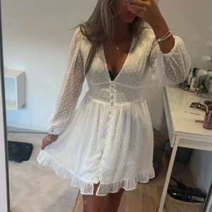 Vit prickig klänning - Charmig vit klänning med prickigt mönster och långa ärmar. Klänningen har en v-ringad design med knappar framtill och en söt volangdetalj vid fållen. Perfekt för en somrig look!