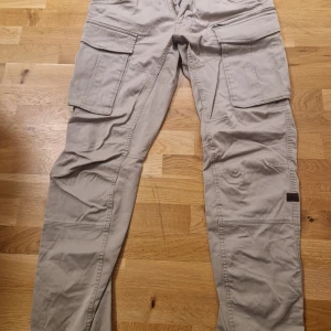 Beige cargopants med fickor G-Star - Säljer ett par beige cargopants med flera praktiska fickor. Byxorna har en straight passform och är perfekta för en avslappnad stil. De är tillverkade i ett slitstarkt material och har knappar och dragkedja framtill.