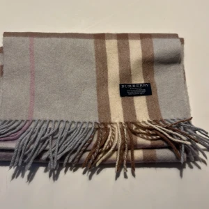 Rutig halsduk från Burberry - Snygg och klassisk rutig halsduk från Burberry i mjuk kashmir. Den har en stilren kombination av beige och grå toner med fransar i ändarna. Perfekt för att hålla dig varm och stilig under kyliga dagar. Den är i nyskick och priset är ej hugget i sten