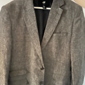 Grå kavaj från H&M - Snygg grå kavaj från H&M i herrstorlek L. Perfekt för både formella och avslappnade tillfällen. Kavajen har en klassisk design med två knappar framtill och fickor. Tillverkad i ett linne material 