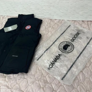 Svart väst från canada goose har alla storlekar  - Säljer en svart dunväst från Canada Goose med knappar och en hög krage. Västen har en broderad logotyp på bröstet och praktiska fickor framtill. Perfekt för kyliga dagar. 
