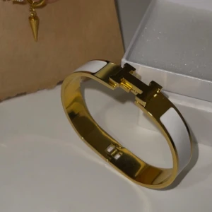Guldfärgad armband med vita detaljer - Helt oanvänd och ny. Materialet är rostfritt stål med 18K guldplätering. 