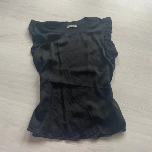 Svart topp med volangdetaljer - Snygg svart topp med volangdetaljer på ärmarna. Tillverkad i Italien och har silke materiel på framsidan.