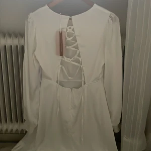 Vit klänning med öppen rygg och snörning - Säljer en elegant vit klänning med långärmad design och en vacker öppen rygg med snörning. Klänningen har en klassisk och stilren look, perfekt för speciella tillfällen. Den är i en kort modell och har en mjuk och flödande passform.