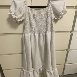 Vit klänning med volanger - Charmig vit klänning med korta puffärmar och volanger i nederkant. Klänningen har en smockad överdel som är utöjbar och är lite onsize i storleken. Lite små okända fläckar(se bild) har använt den vid kanske 3 skolavslutningar men inget utöver det. Det kan säkert gå och tvätta bort