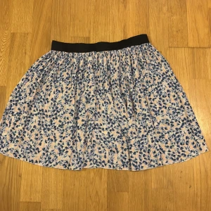 Blommig kjol med svart resår - Söt blommig kjol med blå och vita blommor på en ljus bakgrund. Kjolen har en svart resår i midjan för en bekväm passform. Perfekt för en avslappnad och stilren look.