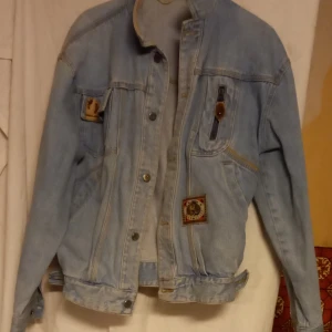 Ljusblå jeansjacka med patchar - Säljer en ljusblå jeansjacka med coola patchar på bröstet. Jackan har knappar framtill och en klassisk krage. Tillverkad i 100% bomull vilket gör den bekväm och hållbar. Perfekt för en avslappnad stil.