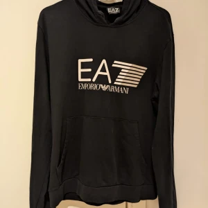 Svart hoodie från Emporio Armani - Snygg svart hoodie från Emporio Armani med stort EA7-tryck på bröstet. Välanvänd men i fint skick. Storlek L, passar M/L. Skickar gärna fler bilder 