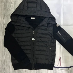 Svart cardigan från Moncler - Säljer en svart jacka från Moncler med huva och dragkedja. Jackan har en quiltad design på framsidan och ribbade ärmslut. Den är långärmad och kombinerar både syntet och stickat material för en unik look. Perfekt för kyligare dagar. I Storlek L perfekt nu inför våren 