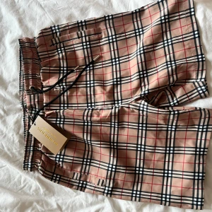 Burberry rutiga shorts med snörning - Snygga shorts från Burberry i klassiskt beige rutigt mönster med svarta och röda linjer. De har elastisk midja med svart snörning och metalländar med Burberry-logga. Perfekta för sommaren och ger en clean, lyxig vibe.