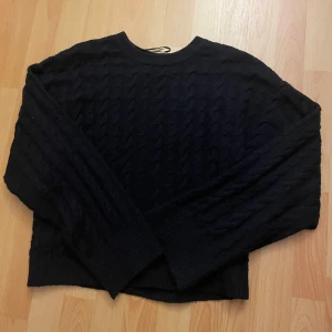 Svart kabelstickad tröja - Mysig svart kabelstickad tröja med rund hals och långa ärmar. Tröjan har breda ribbade muddar vid ärmslut och nederkant, vilket ger en snygg och avslappnad look. Perfekt för dig som gillar klassisk stil med en twist.