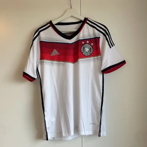 Tysklands matchtröja Adidas S - Säljer en officiell Tyskland fotbollströja från Adidas i storlek S. Tröjan är vit med svarta och röda detaljer, har V-ringning och korta ärmar. Klassiska Adidas-ränder på axlarna och broderat landslagsmärke på bröstet. Tillverkad i lätt och ventilerande polyester.