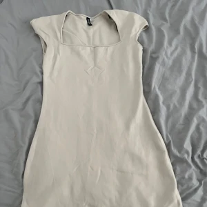 Beige kort klänning från Collusion - Snygg beige kortklänning från Collusion med fyrkantig ringning och ärmlös design. Klänningen har en liten slits framtill och är gjord i mjuk bomull. Perfekt för dig som gillar minimalistisk och clean stil.