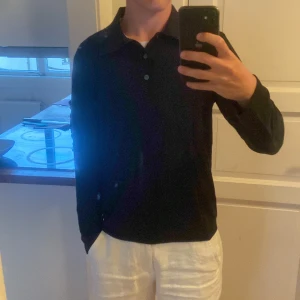 Svart långärmad pikétröja i bomull - Säljer en svart långärmad pikétröja med klassisk krage och knäppning framtill. Tröjan är i mjuk bomull och har en clean, stilren look som funkar till många outfits. Perfekt för dig som gillar enkel och tidlös stil.