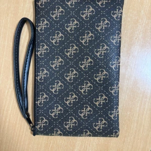 Svart kuvertväska med mönster - Stilren svart kuvertväska med beige mönster och handledsrem. Väskan har dragkedja upptill och är tillverkad i syntetmaterial med ett elegant tryckt mönster över hela ytan. Perfekt för att bära det viktigaste och ger en snygg touch till din outfit.