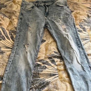 Ljusblå slitna jeans med raka ben - Säljer ett par ljusblå jeans med snygga slitningar och raka ben. Jeansen har klassisk femficksdesign, dragkedja och bälteshällor. De är tillverkade i denim och har en tvättad look som ger en cool vibe.