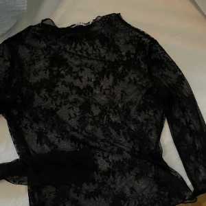 Svart spetsblus från NA-KD - Säljer en transparent svart spetsblus från NA-KD i storlek S. Blusen har lång ärm och är täckt av ett blommigt spetsmönster som ger en riktigt snygg och edgy vibe. Perfekt att styla lager på lager eller med ett linne under.