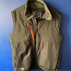 Grön väst från Helly Hansen Workwear - Grön väst från Helly Hansen Workwear med hög krage och hel dragkedja framtill. Västen har en orange dragkedja på bröstfickan och två sidofickor. Materialet är slitstarkt och passar perfekt för utomhusaktiviteter eller jobb. Diskret HH-logga nertill.